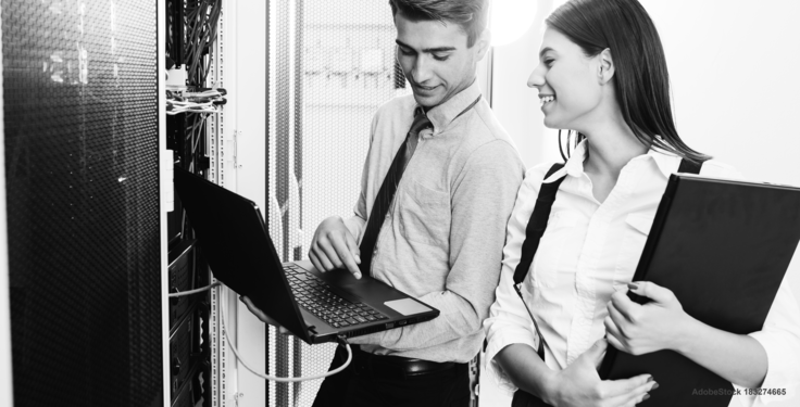 Symbolbild Ein Mann und eine Frau stehen vor einem Serverschrank und schauen auf einen Laptop